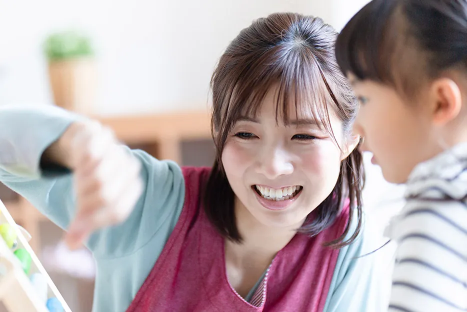 エプロン姿の保育士が、女の子と一緒に笑顔でおもちゃを指さしている