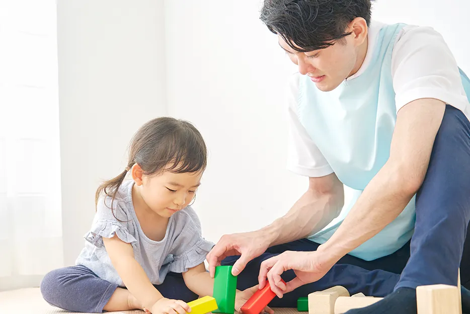 男性保育士が、積み木で遊ぶ女の子をそばで見守っている