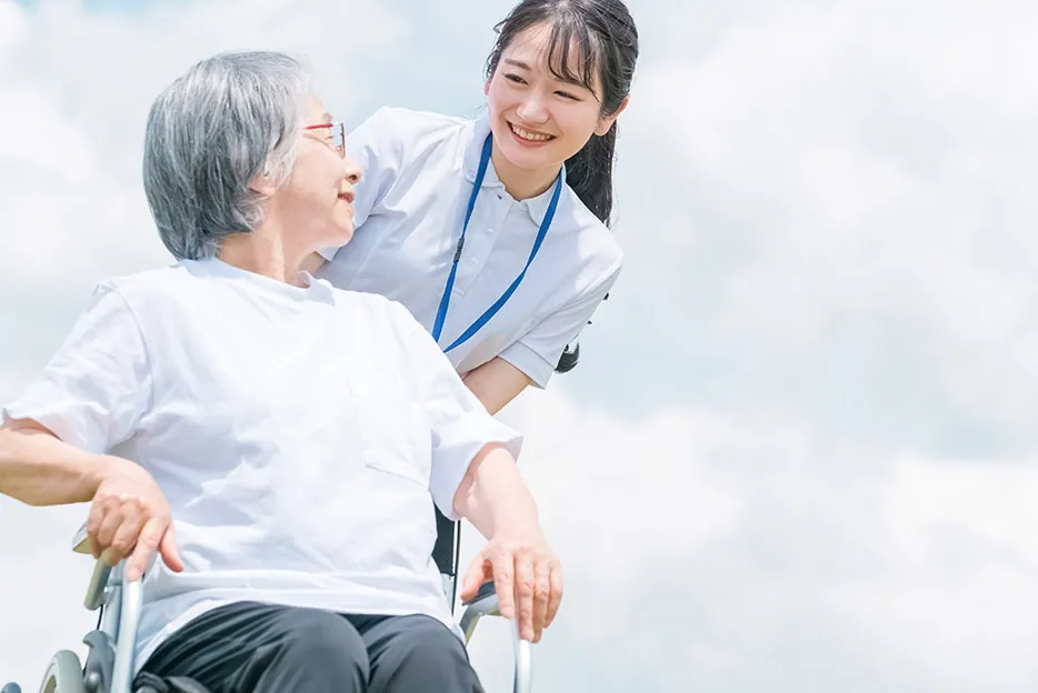 青空の下、車いすの女性と若い職員が笑顔で見つめ合っている