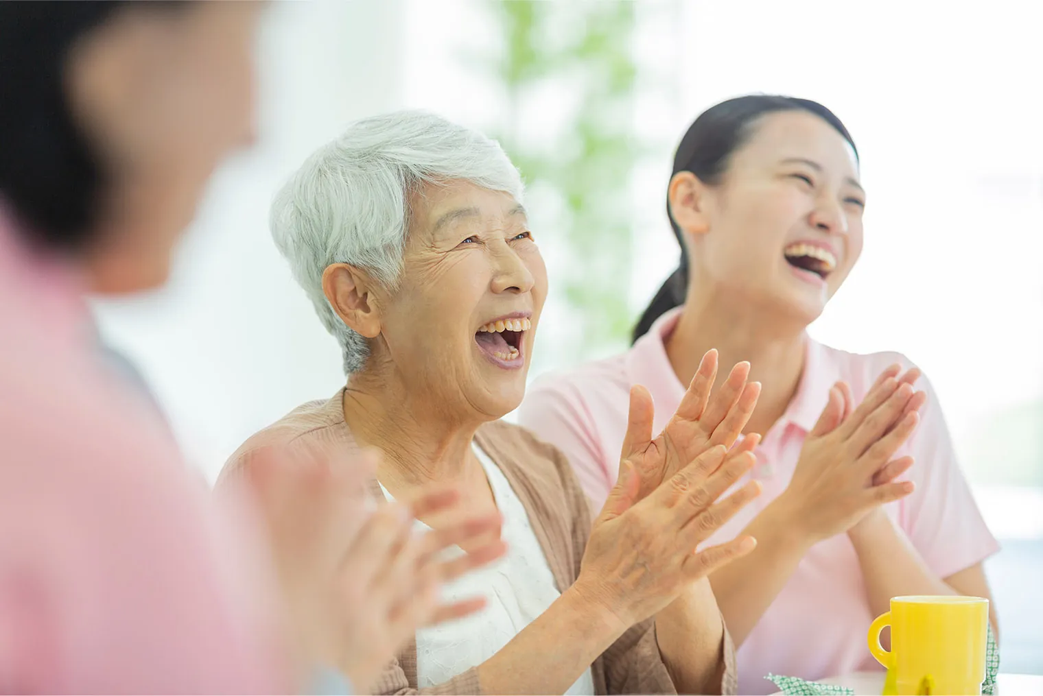 デイサービスで高齢の女性と若い職員が声をあげて笑いながら拍手している
